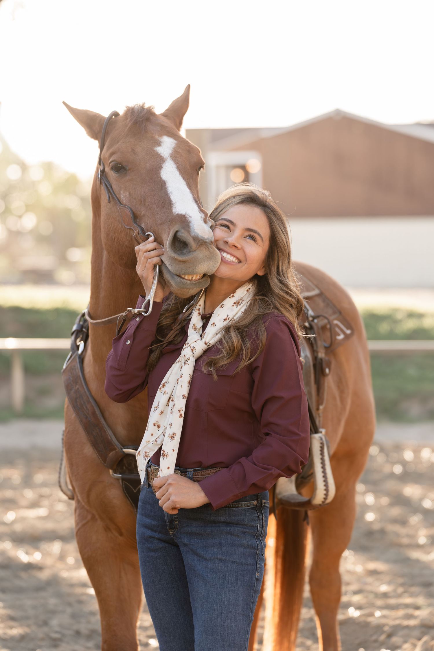 Smiling woman in burgundy shirt and scarf stands beside a chestnut horse at a sunny ranch.
