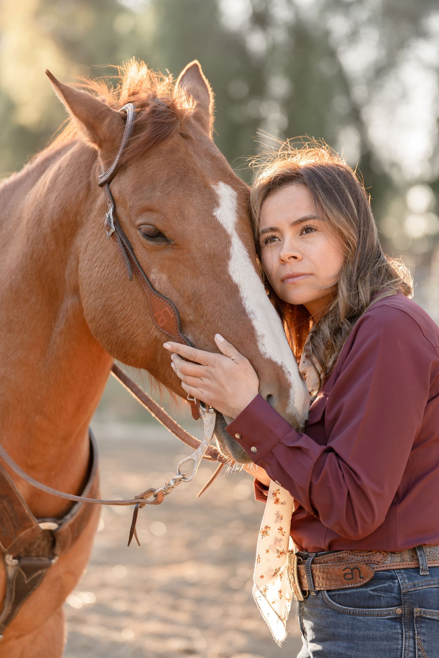Young woman in red shirt gently holds a chestnut horse with white blaze in warm golden sunlight outdoors.