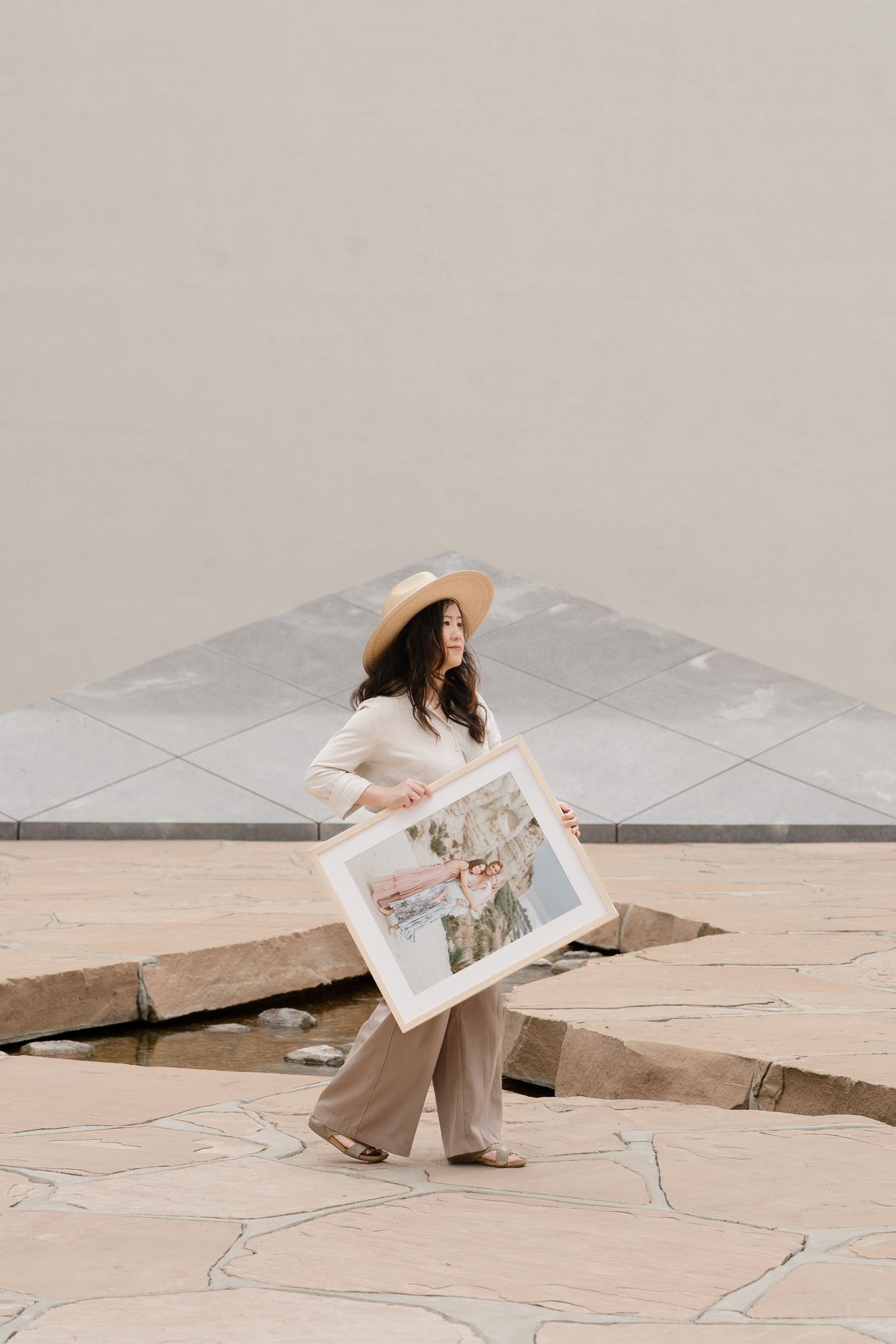 Woman in wide-brim hat and wide-leg pants holds framed artwork outdoors on stone plaza.
