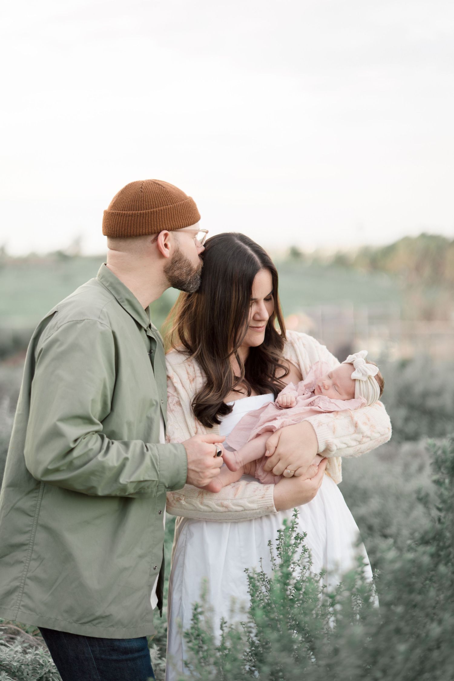 New parents share a tender moment as father kisses mother holding newborn baby in pink outdoors.