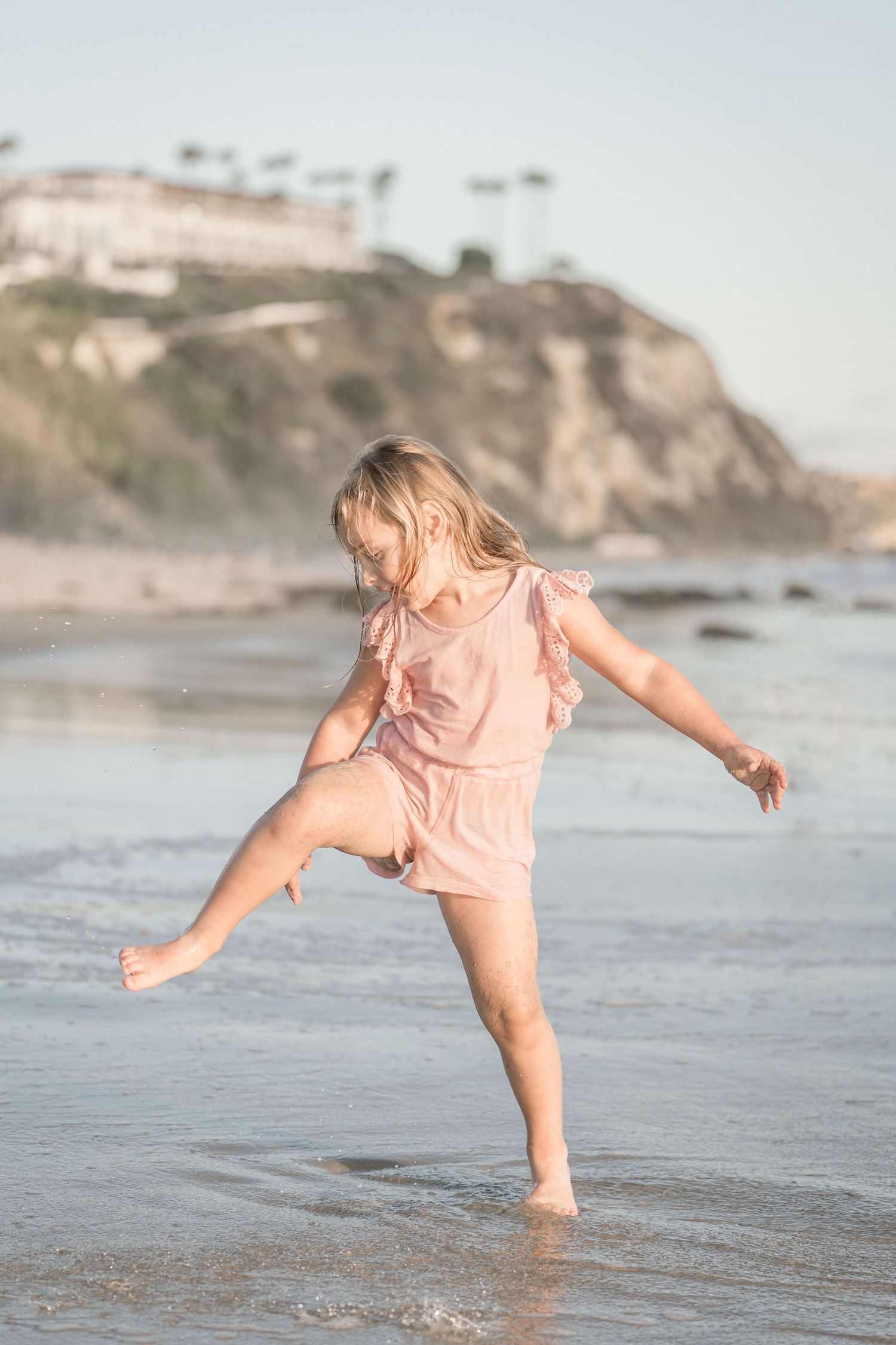 Young blonde girl in pink romper joyfully kicks water at the beach during golden hour.