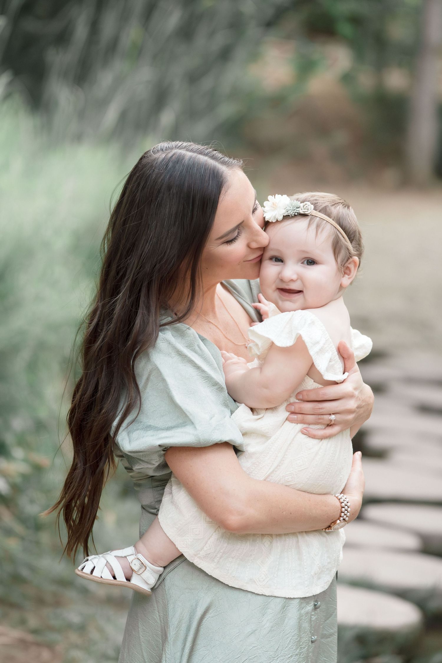 Loving mother with long dark hair kisses happy baby girl in white dress during outdoor family photo session.