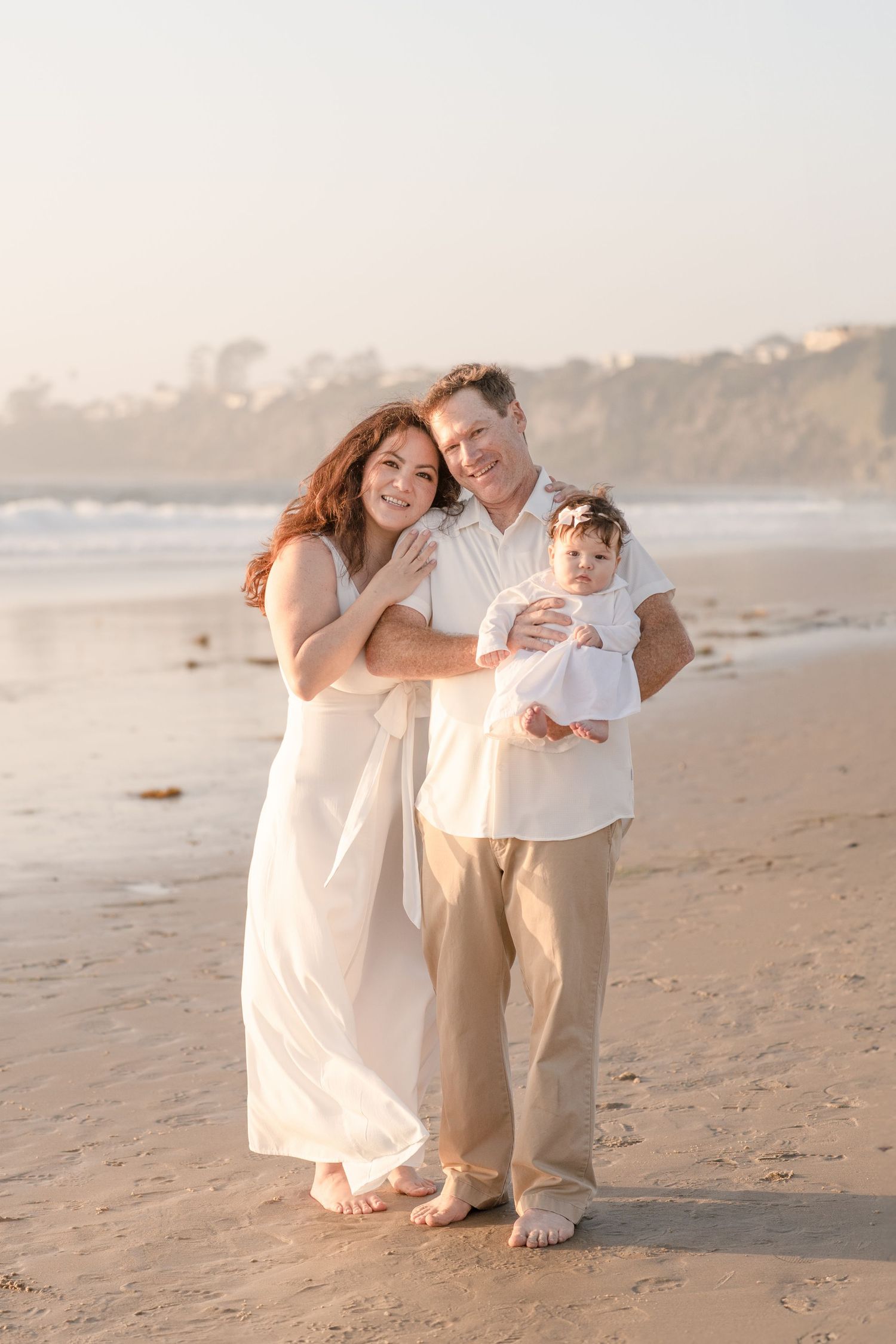 A happy family of three poses barefoot on a sandy beach at golden hour, parents holding their baby in white outfits.