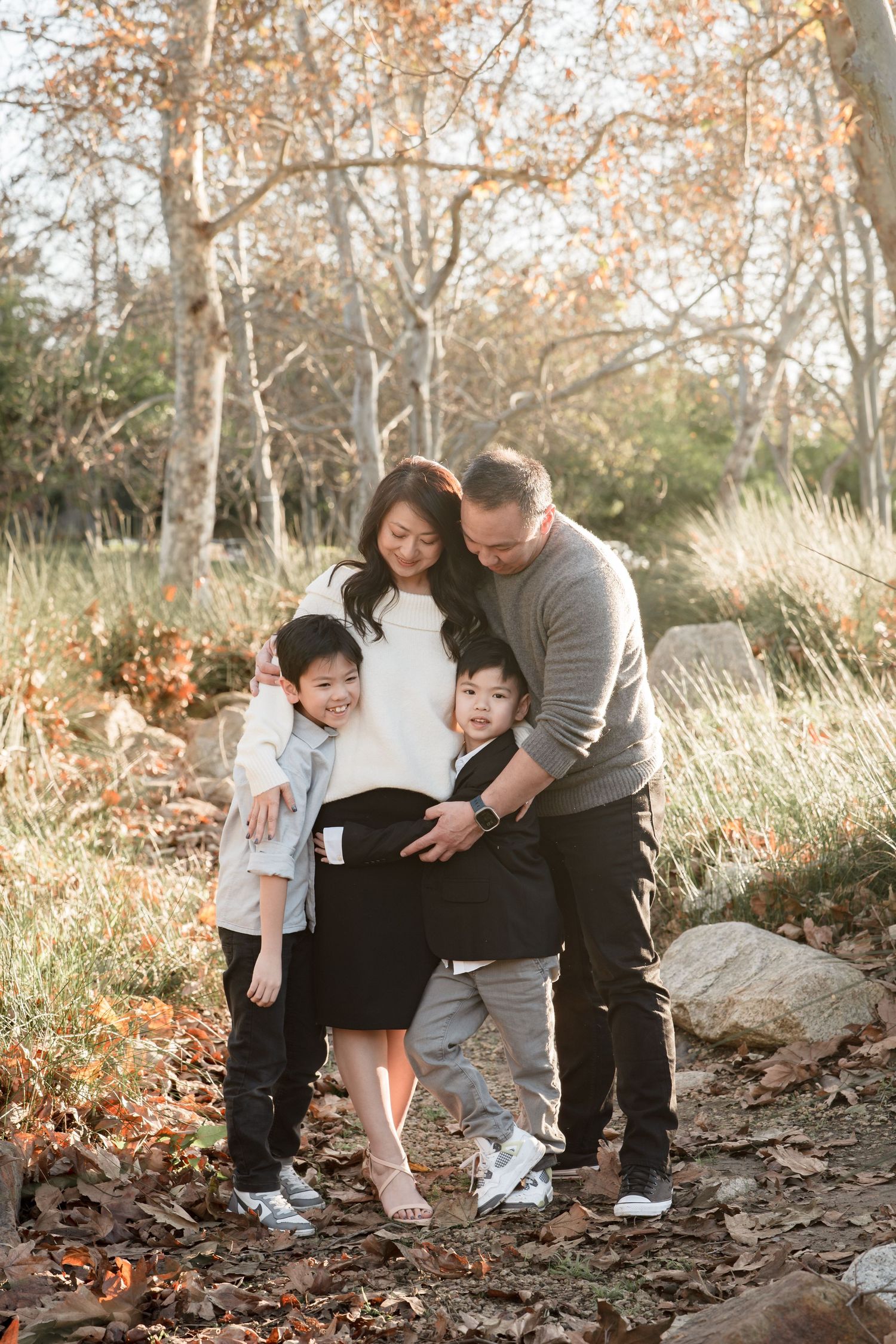 Family of four posing outdoors in autumn leaves, parents embracing two young children among bare trees in golden light.