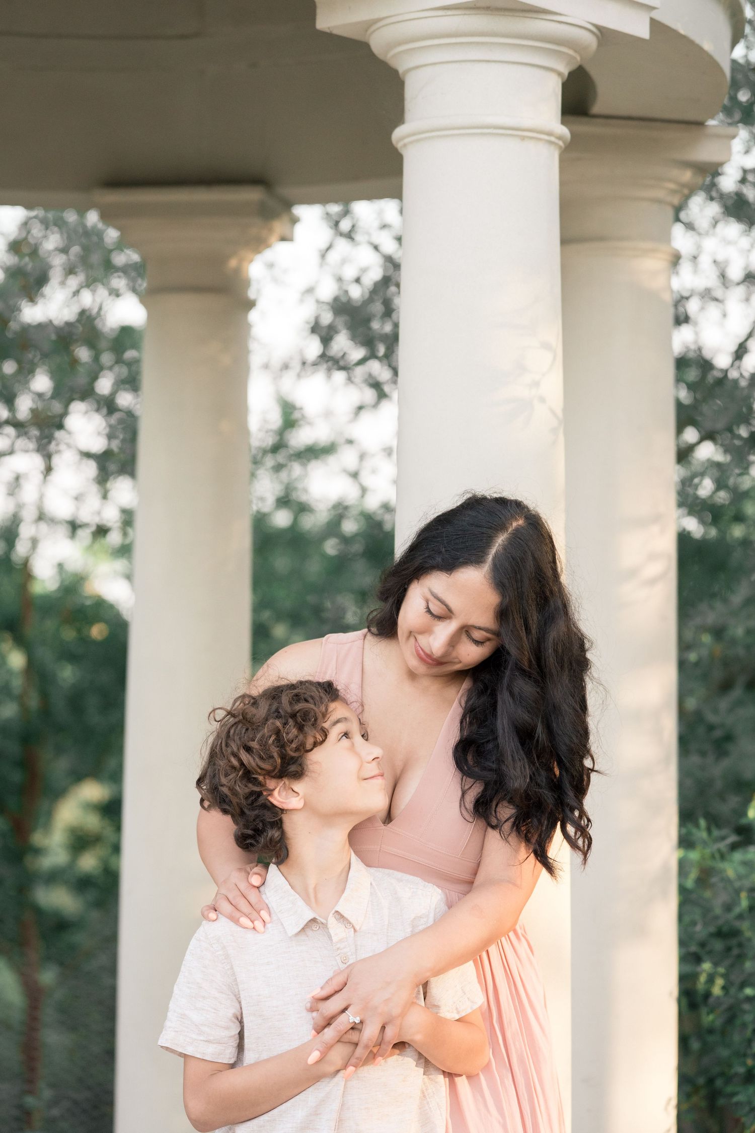 A mother in a pink dress tenderly embraces her curly-haired son between white columns in a garden.