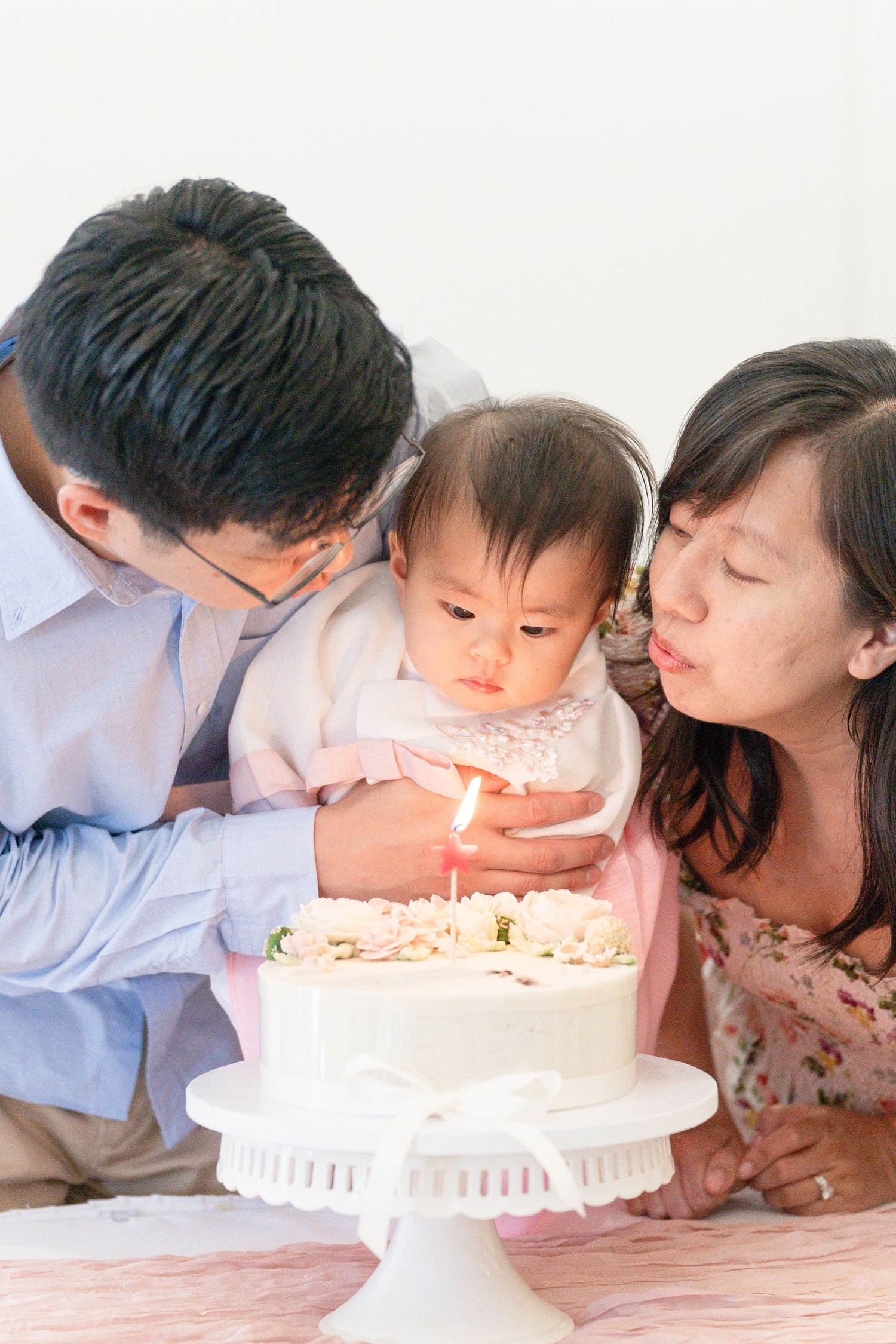 Parents help their baby girl blow out a candle on a white floral birthday cake during a first birthday celebration.