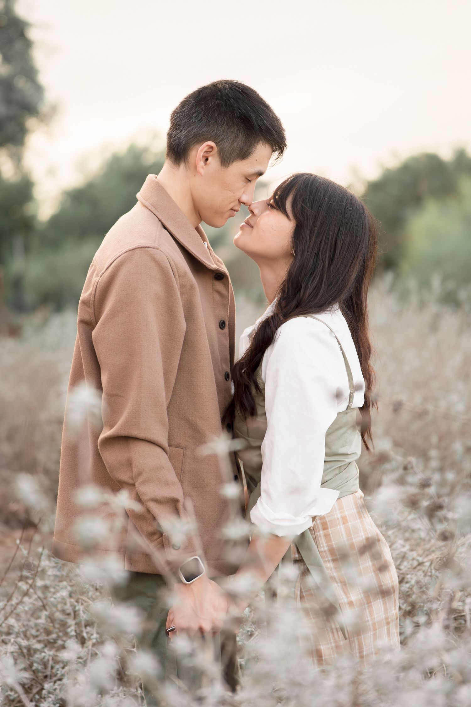 Couple shares an intimate almost-kiss moment surrounded by soft white wildflowers in a dreamy field.