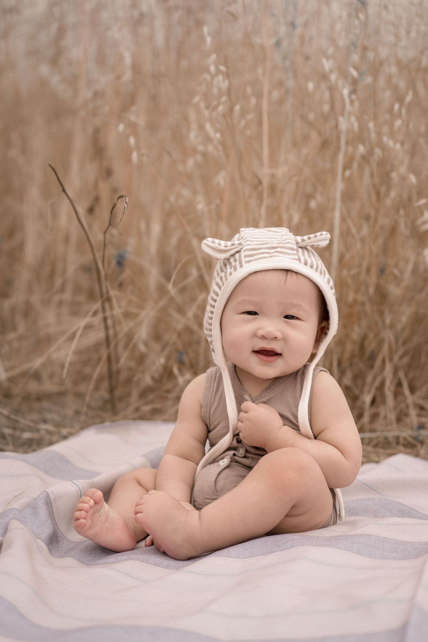 An adorable smiling baby sits on a blanket wearing a knit bonnet with ears in front of golden tall grass.