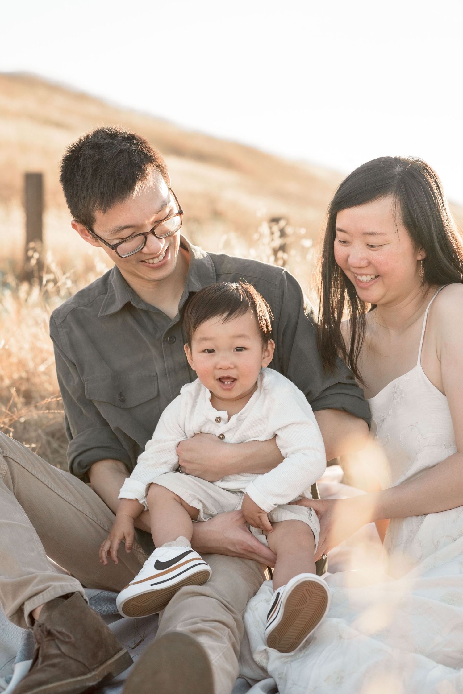 Happy Asian family sits together in golden field, parents smiling at laughing baby in white outfit.