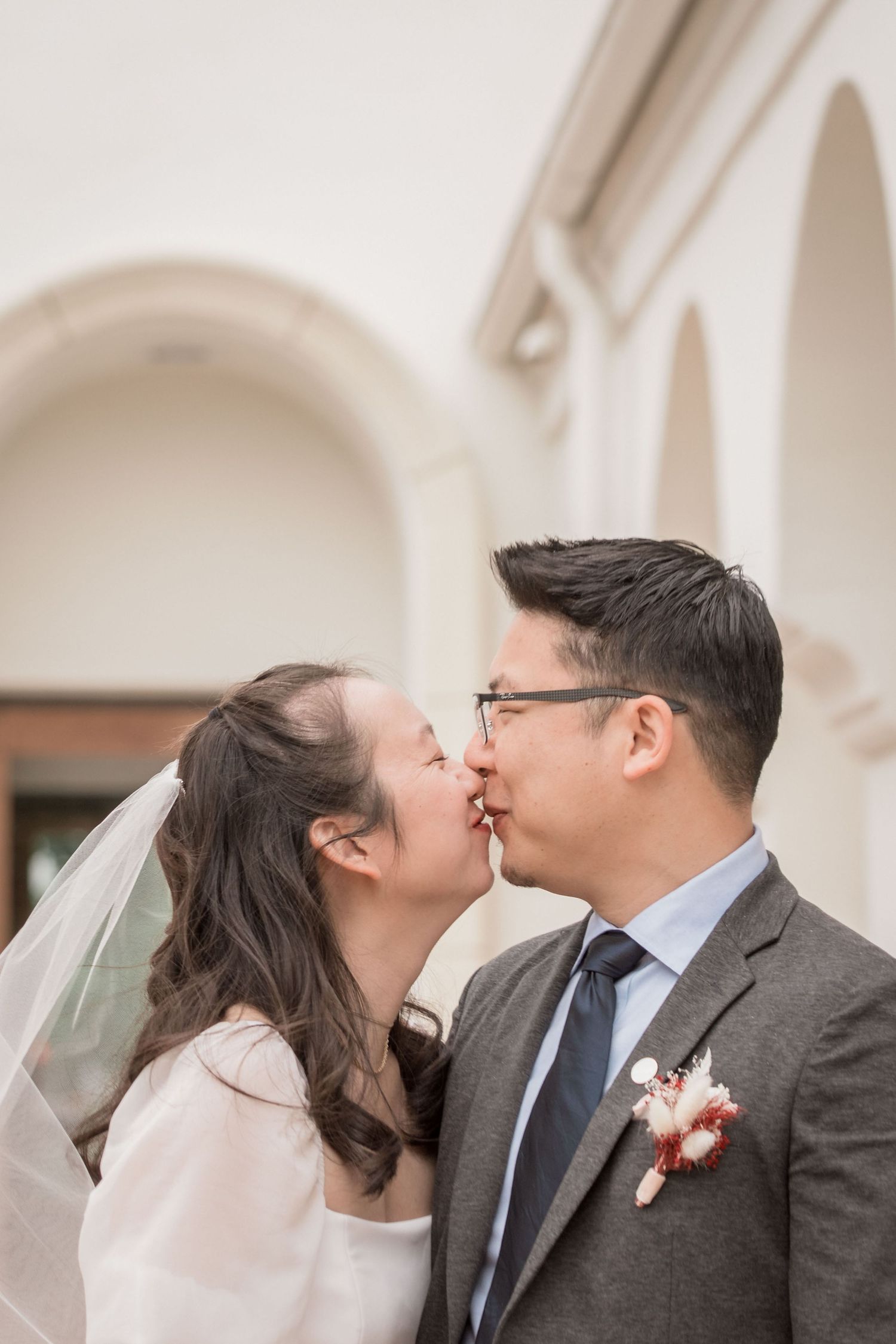 Asian bride and groom share a tender kiss near white arched architecture.