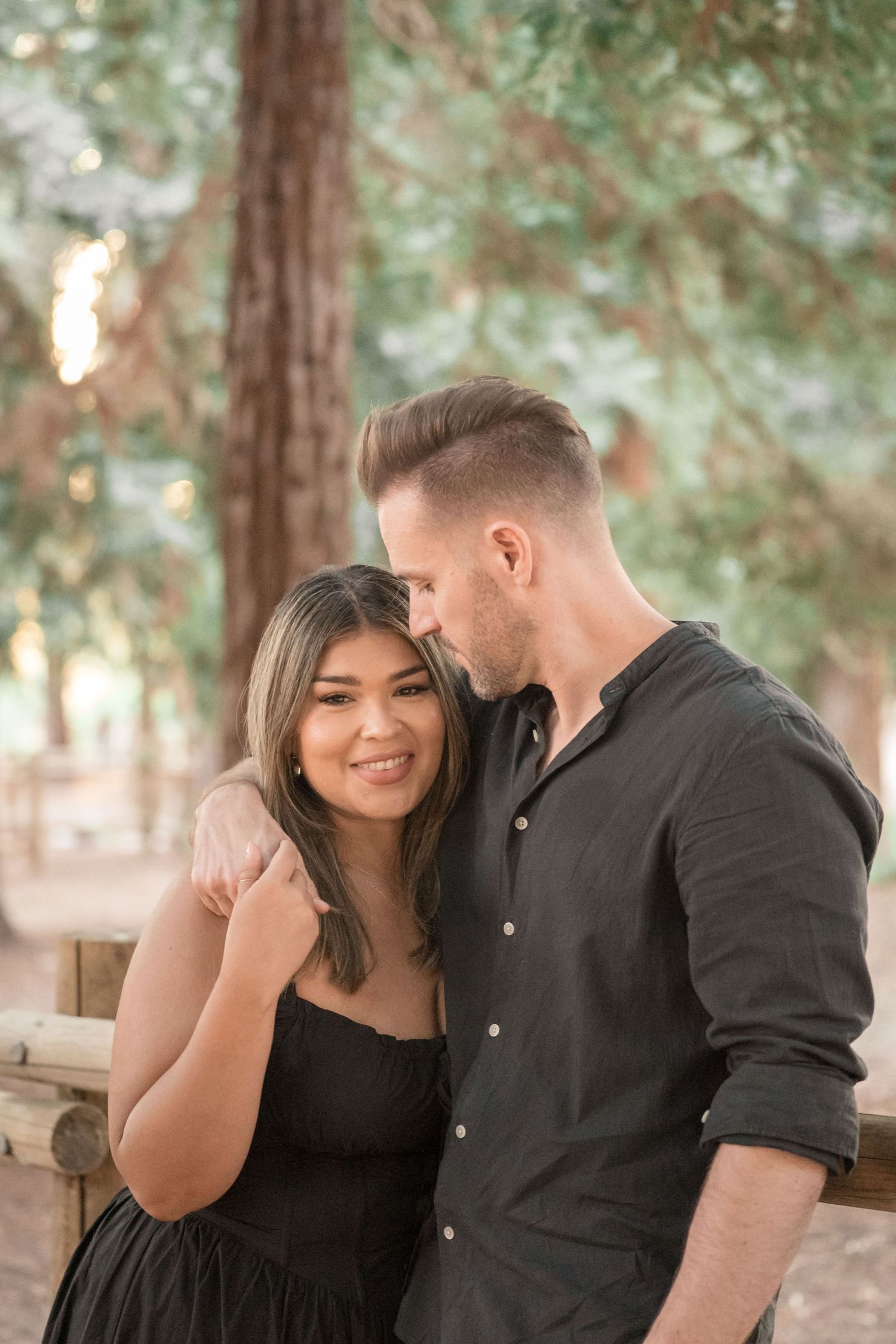 Couple embracing outdoors in a forest setting, woman in black dress smiling warmly at camera.