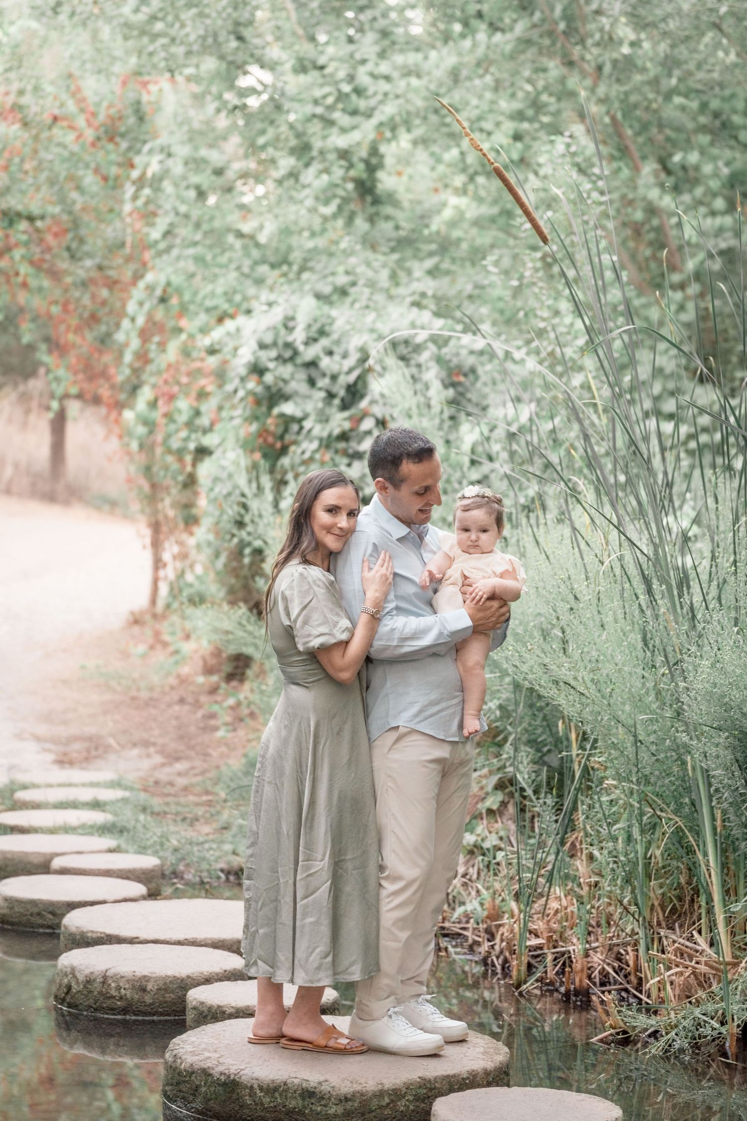 Young family of three standing on garden stepping stones, parents holding infant in green setting.