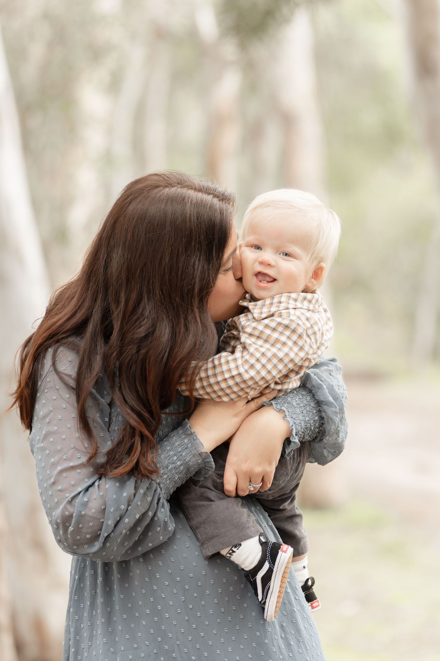 Brunette mother tenderly kisses her smiling blonde baby boy while holding him in a wooded setting.