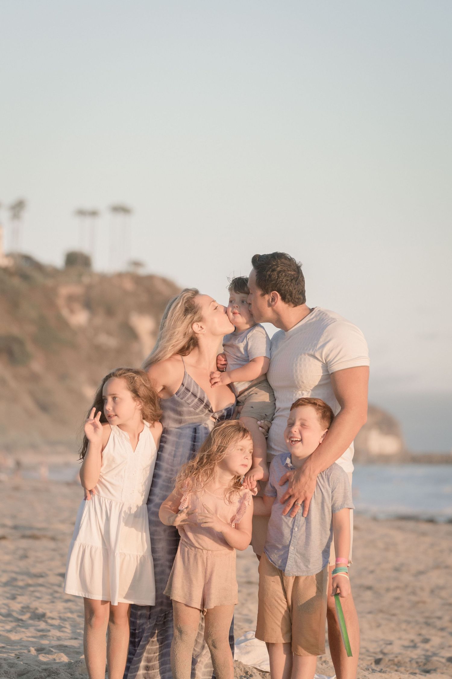 Family of five standing on beach at sunset, parents kissing while holding toddler.