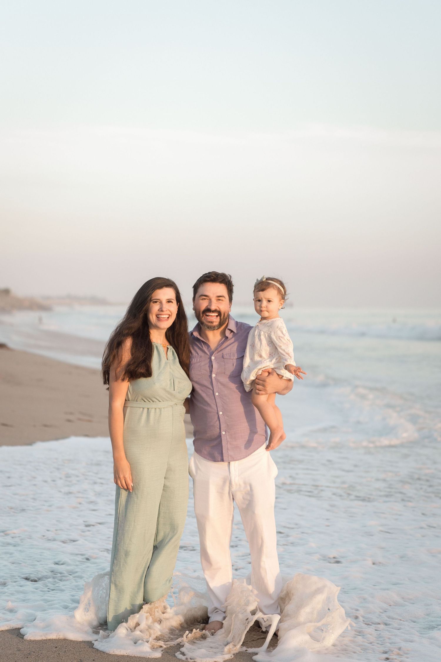 Happy family with bearded dad holding toddler and mom in green jumpsuit standing on beach at sunset.