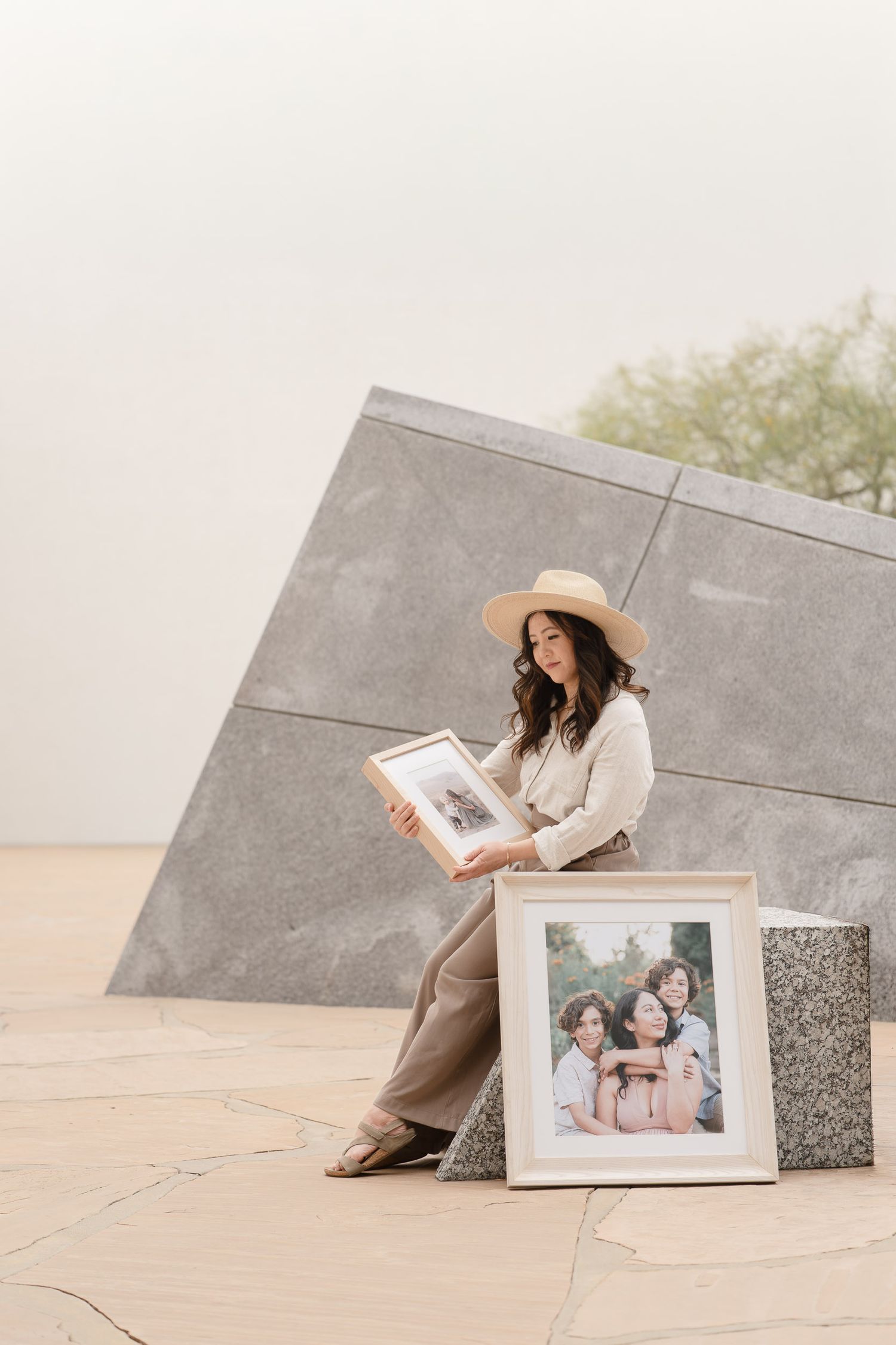 Woman in straw hat sits outdoors holding a framed photo beside a large portrait of two women smiling.
