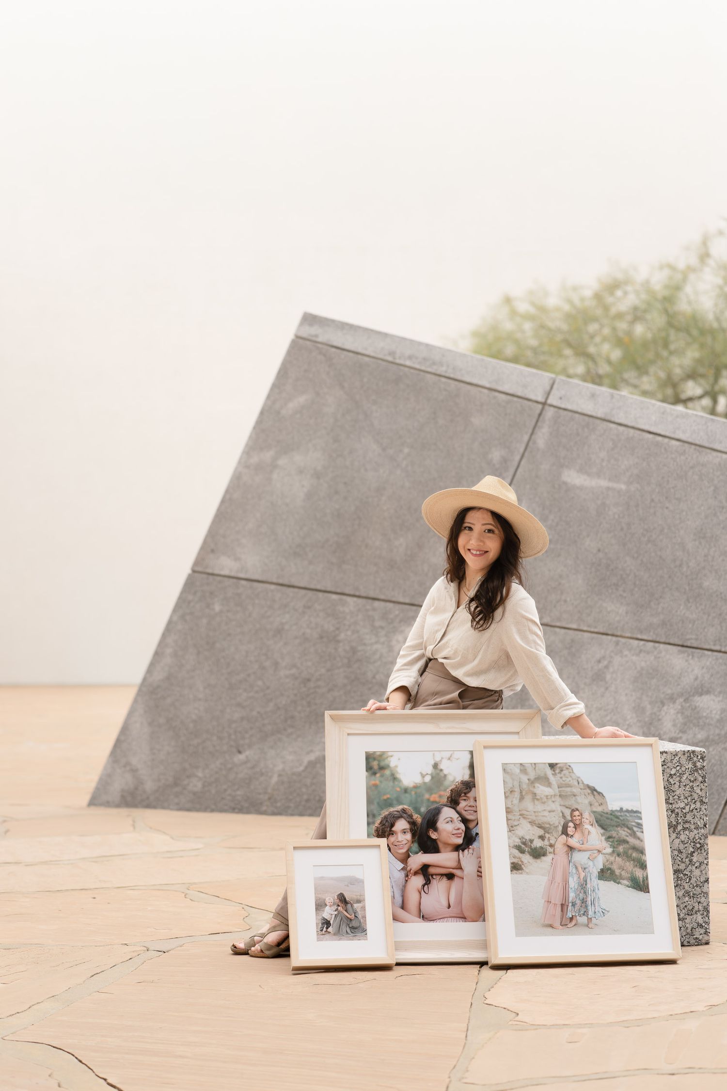 Woman in beige outfit and hat poses outdoors with framed family portrait photographs displayed on stone surface.