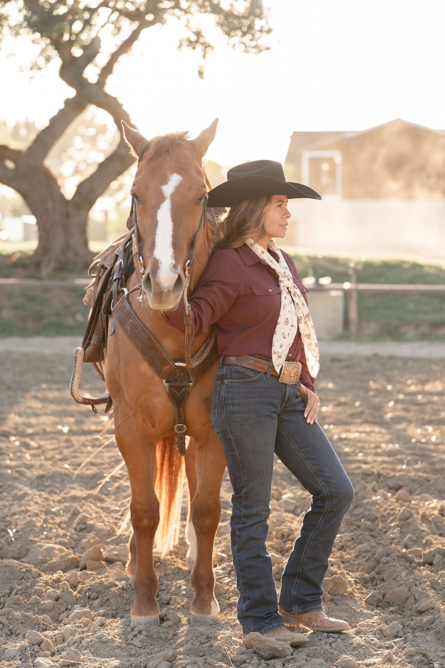 A cowgirl in a black hat and jeans stands beside a chestnut horse in a sandy paddock at golden hour.