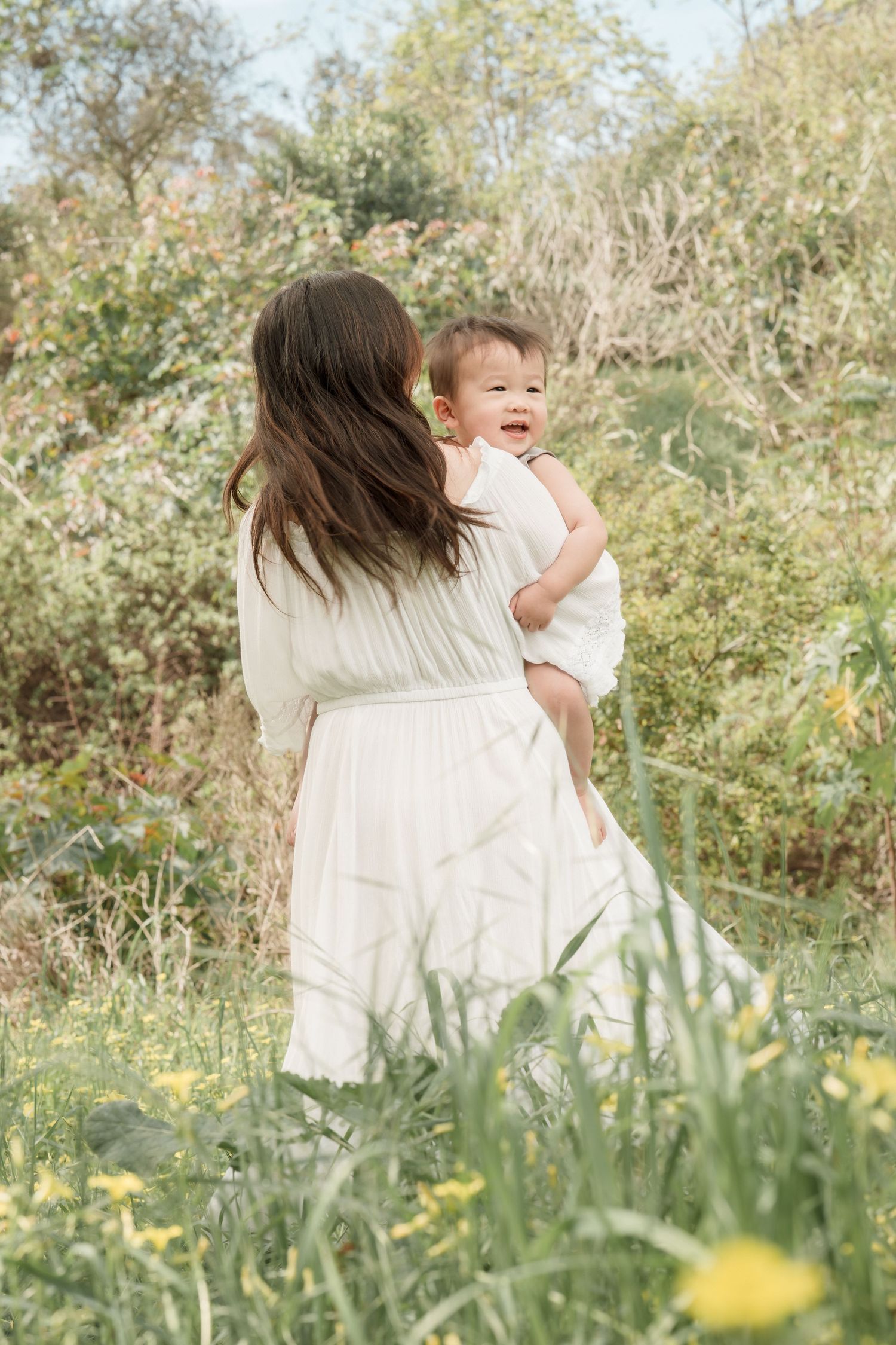 A mother in a white dress holds her laughing baby while standing among lush green bushes in a sunny outdoor setting.