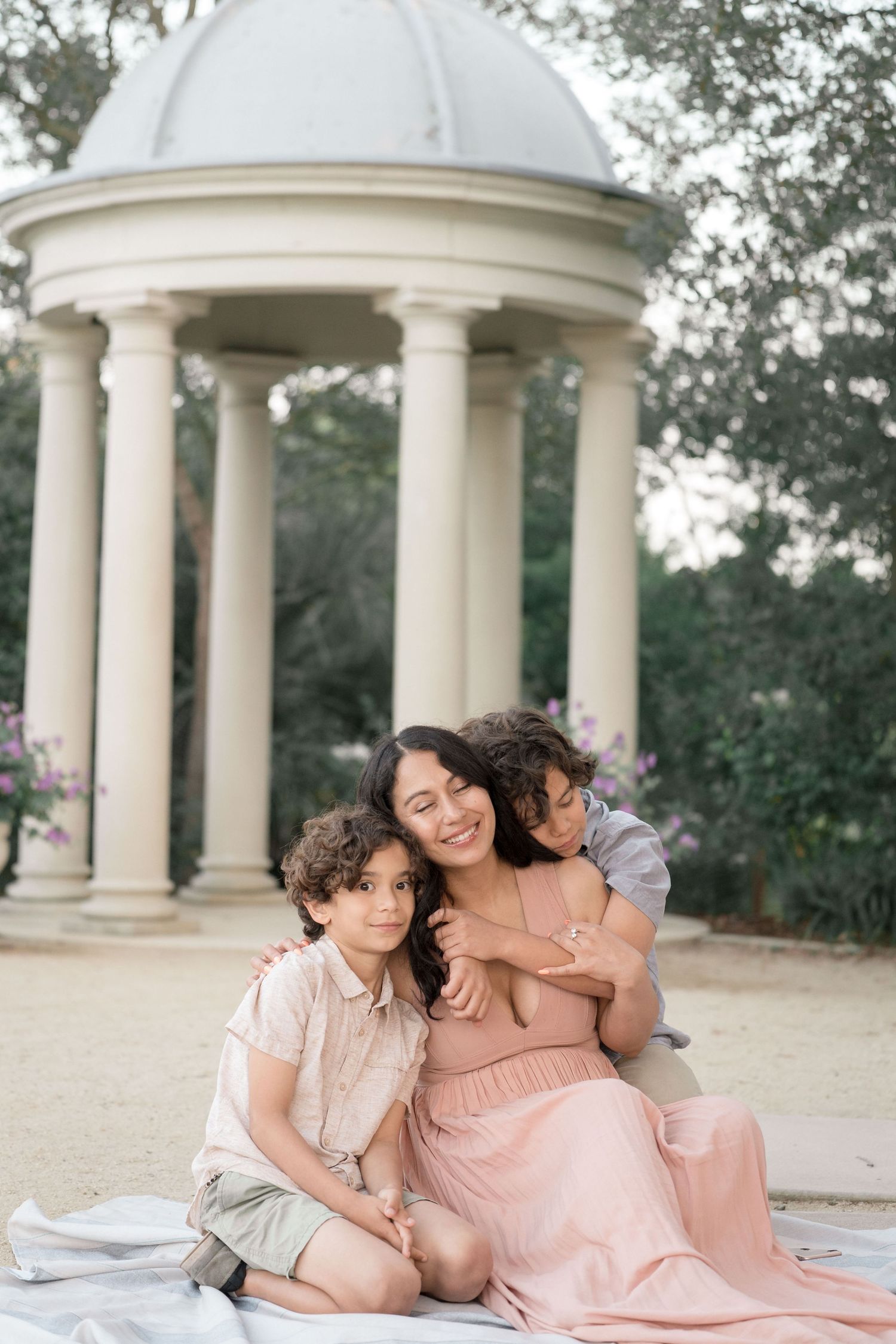 A smiling mother sits with her two young sons on a blanket near a white garden gazebo.