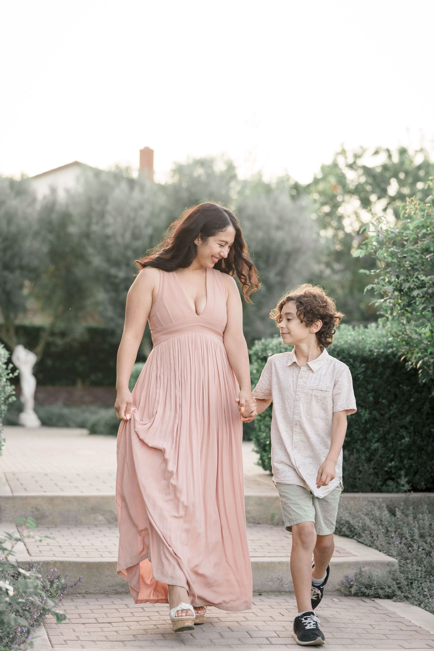 Smiling mother in blush maxi dress walks hand in hand with young son outdoors.