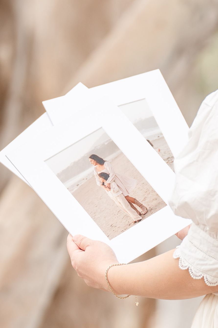 Hands holding printed beach maternity photos with white borders in soft natural light.