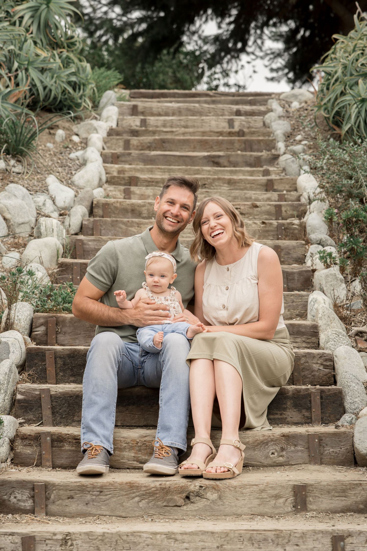 Happy family with baby sitting on rustic wooden steps surrounded by lush greenery outdoors.