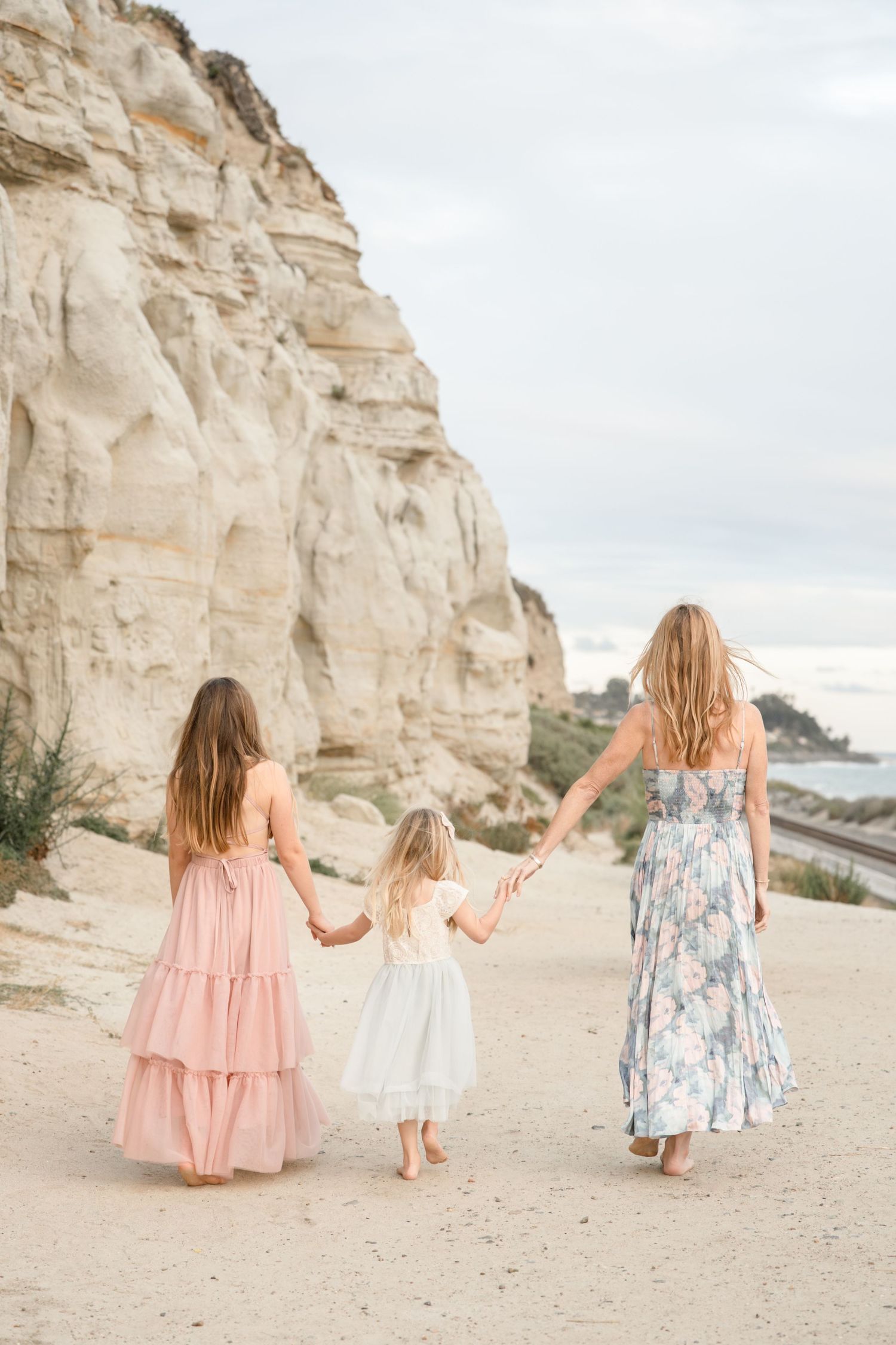 Three girls in maxi dresses hold hands walking on sandy beach near white cliffs.