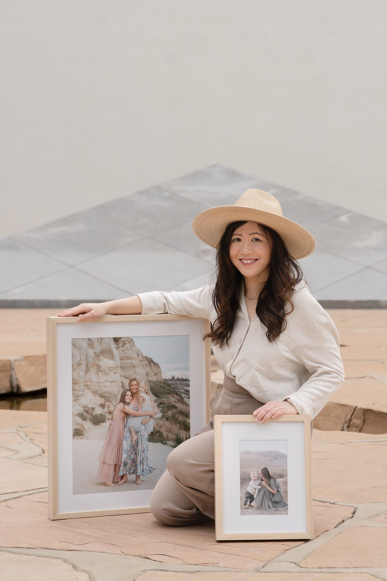 Woman in beige hat sitting on stone floor displaying two framed portrait photographs outdoors.