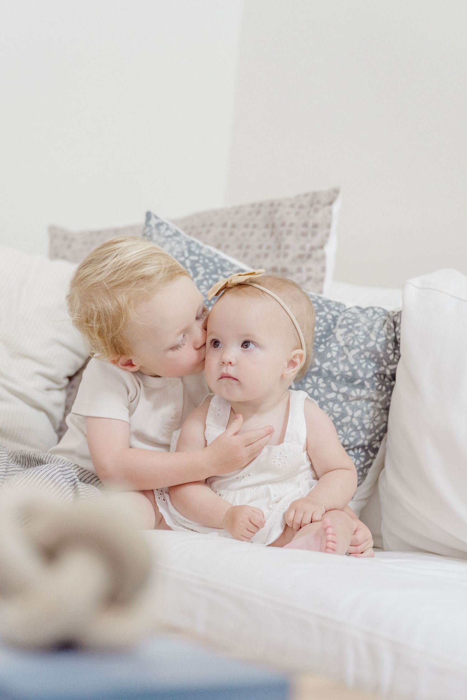 Blond toddler boy gently kissing baby girl with gold headband seated on white cushioned sofa.