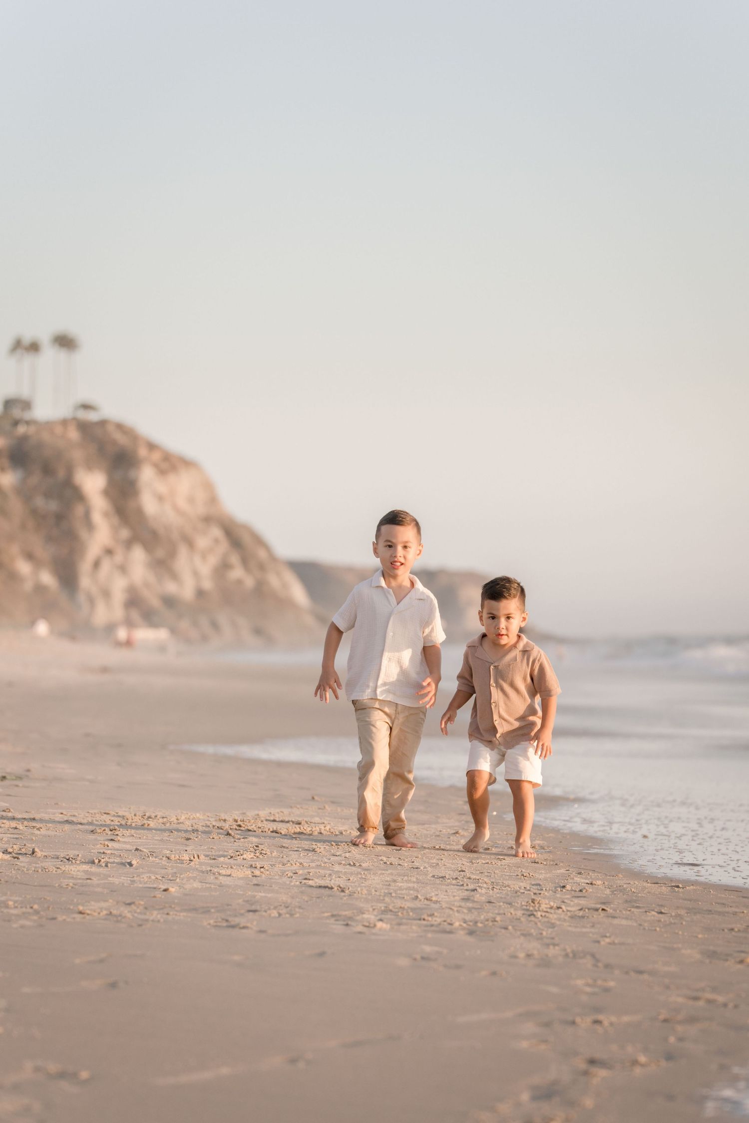 Two young brothers in neutral outfits running barefoot along a sandy beach at golden hour.