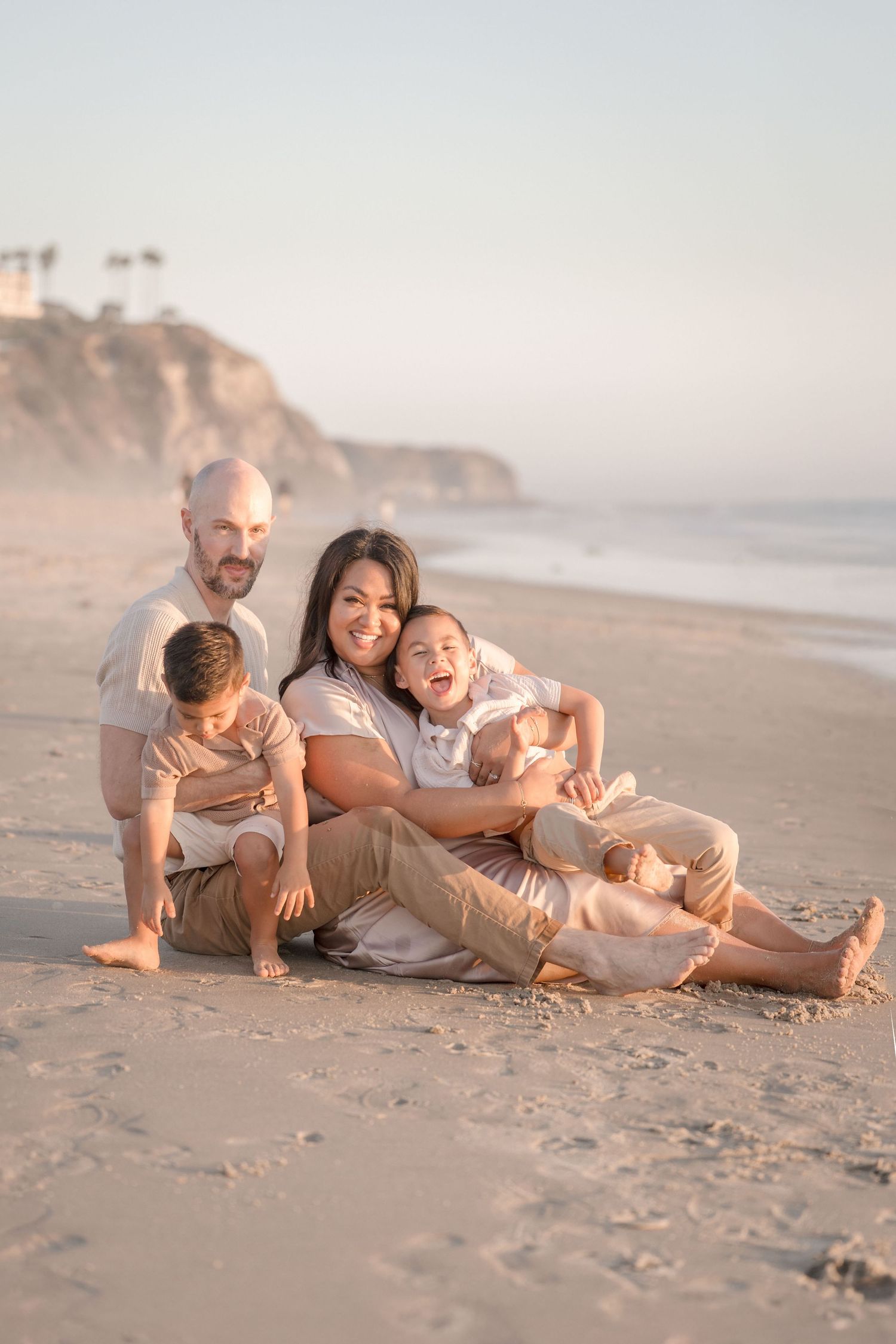 A joyful family of four sits together on a sandy beach, smiling and laughing during a golden hour portrait session.