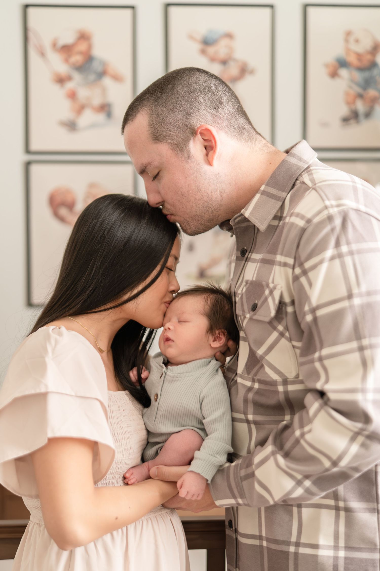 Loving parents kissing over sleeping newborn baby in nursery with vintage bear illustrations on wall.