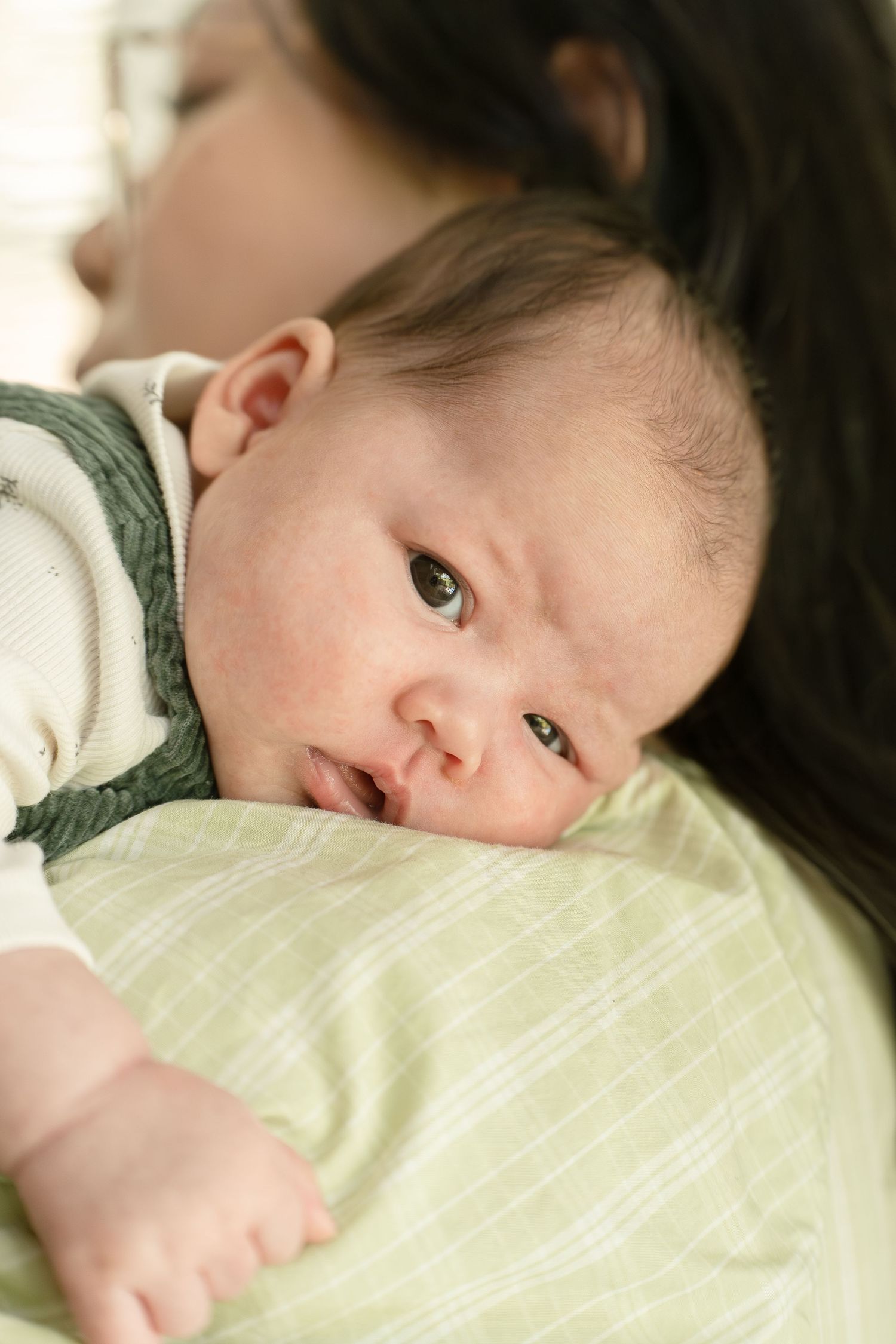 Close-up portrait of alert newborn baby with dark hair resting on mothers shoulder in green striped cloth.