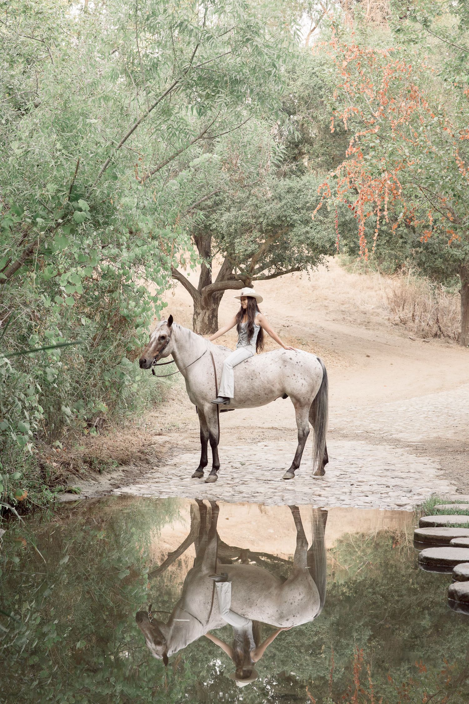 Woman in white outfit and hat riding a white horse near a reflective pool surrounded by lush green trees.