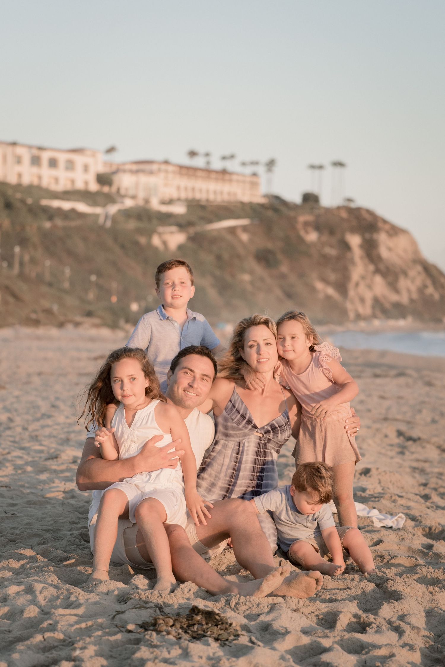 Happy family of six sitting together on a sandy beach at golden hour with coastal cliffs behind.