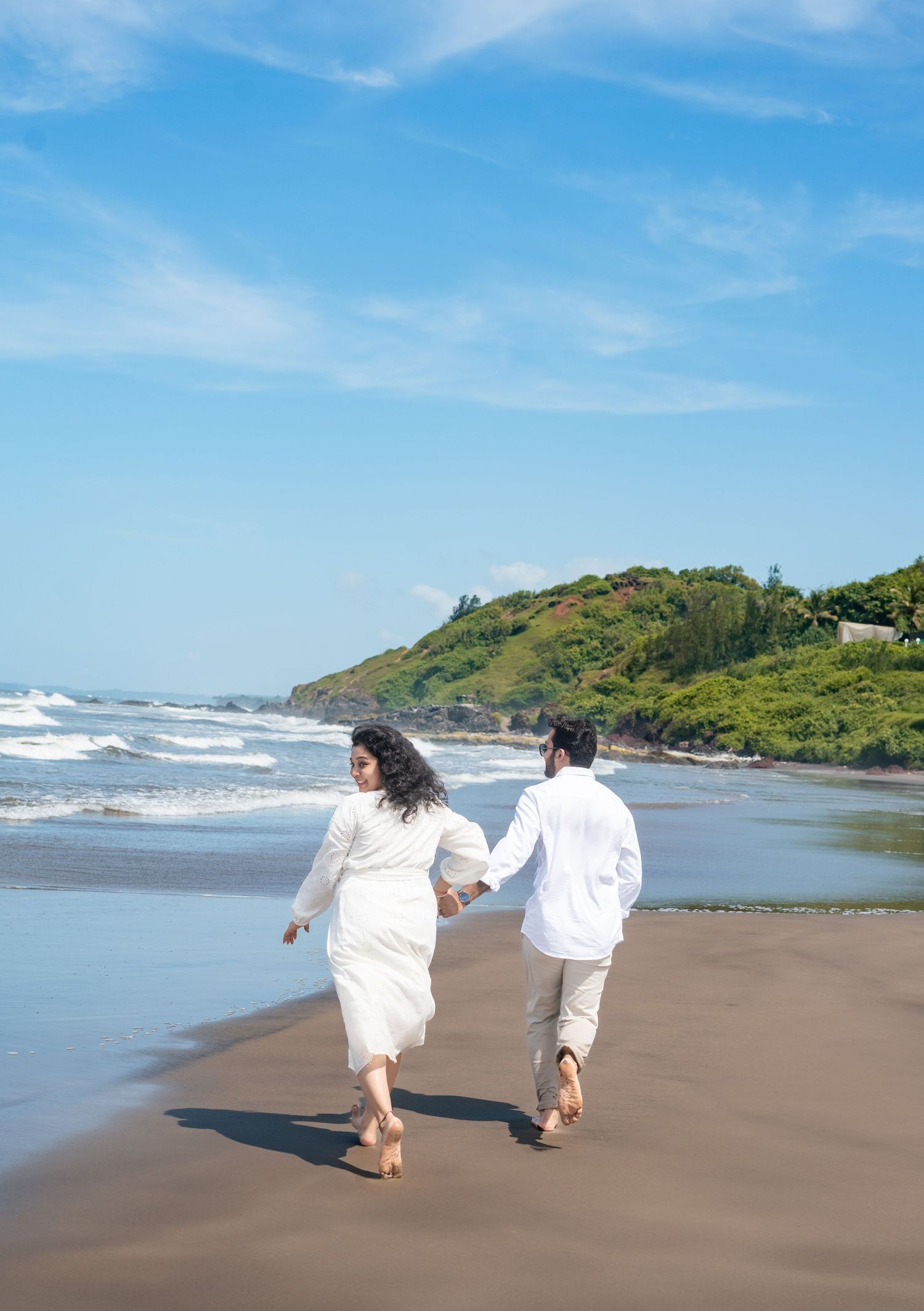Two people walking hand in hand along a scenic beach with mountains in the background.