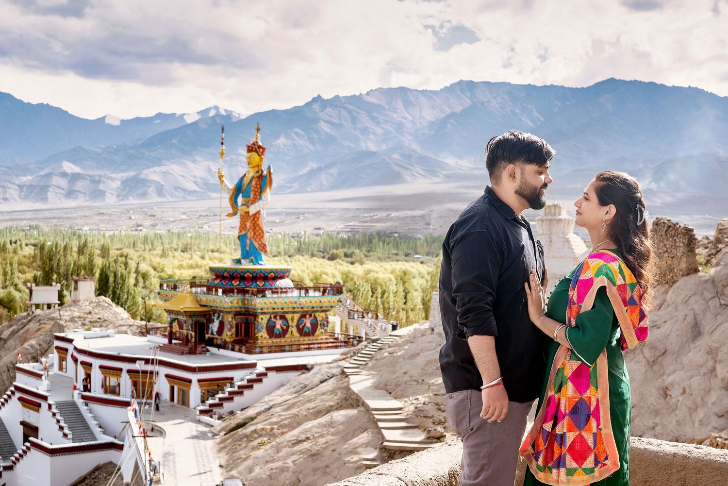 A couple poses against dramatic mountain landscapes in Ladakh with traditional architecture visible.