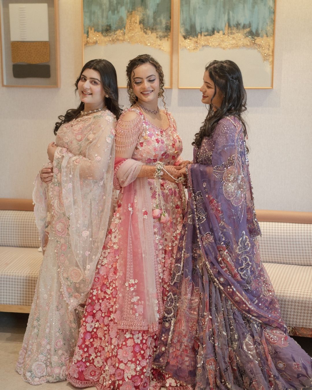 Three women in elegant embroidered Indian lehengas in white, pink, and purple pose together at a festive celebration.