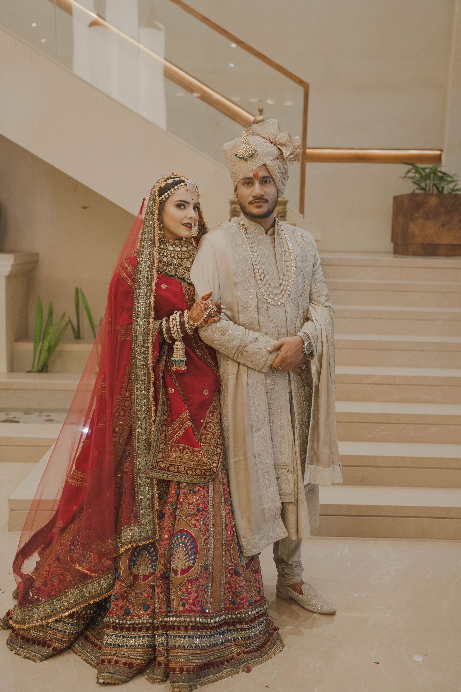 Indian bride in red lehenga and groom in ivory sherwani pose together on wedding day near staircase.