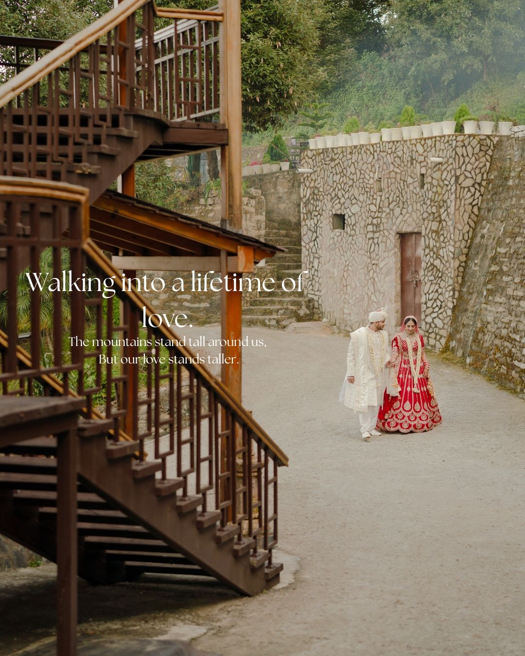 A bride in red lehenga and groom in white sherwani walk together near stone steps and walls in a mountain wedding setting.