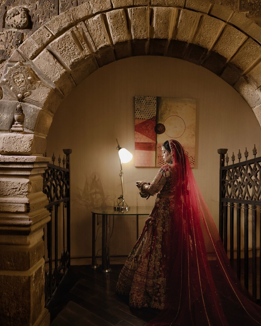 A bride in a red lehenga and dupatta stands under a stone arch in a warmly lit heritage venue.