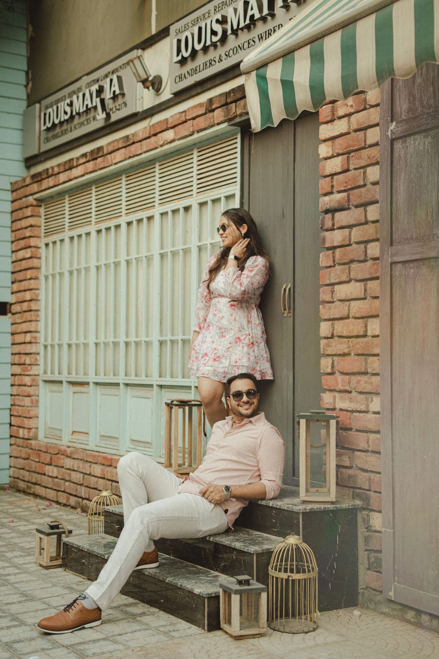 Couple posing outside a vintage brick building with bird cages, woman in floral dress standing behind seated man in pink shirt.