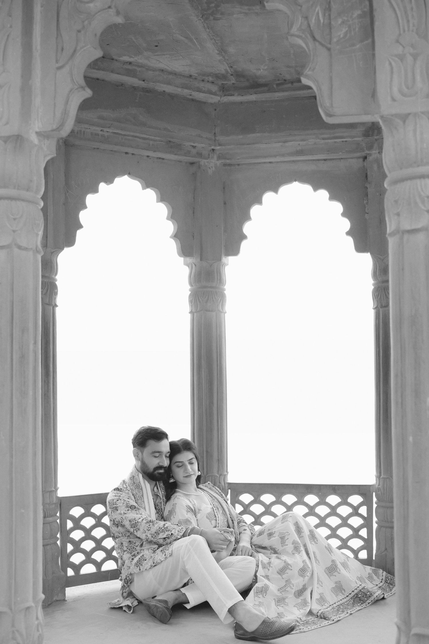Black and white photo of a couple in traditional Indian attire sitting together in ornate Mughal arch alcove.