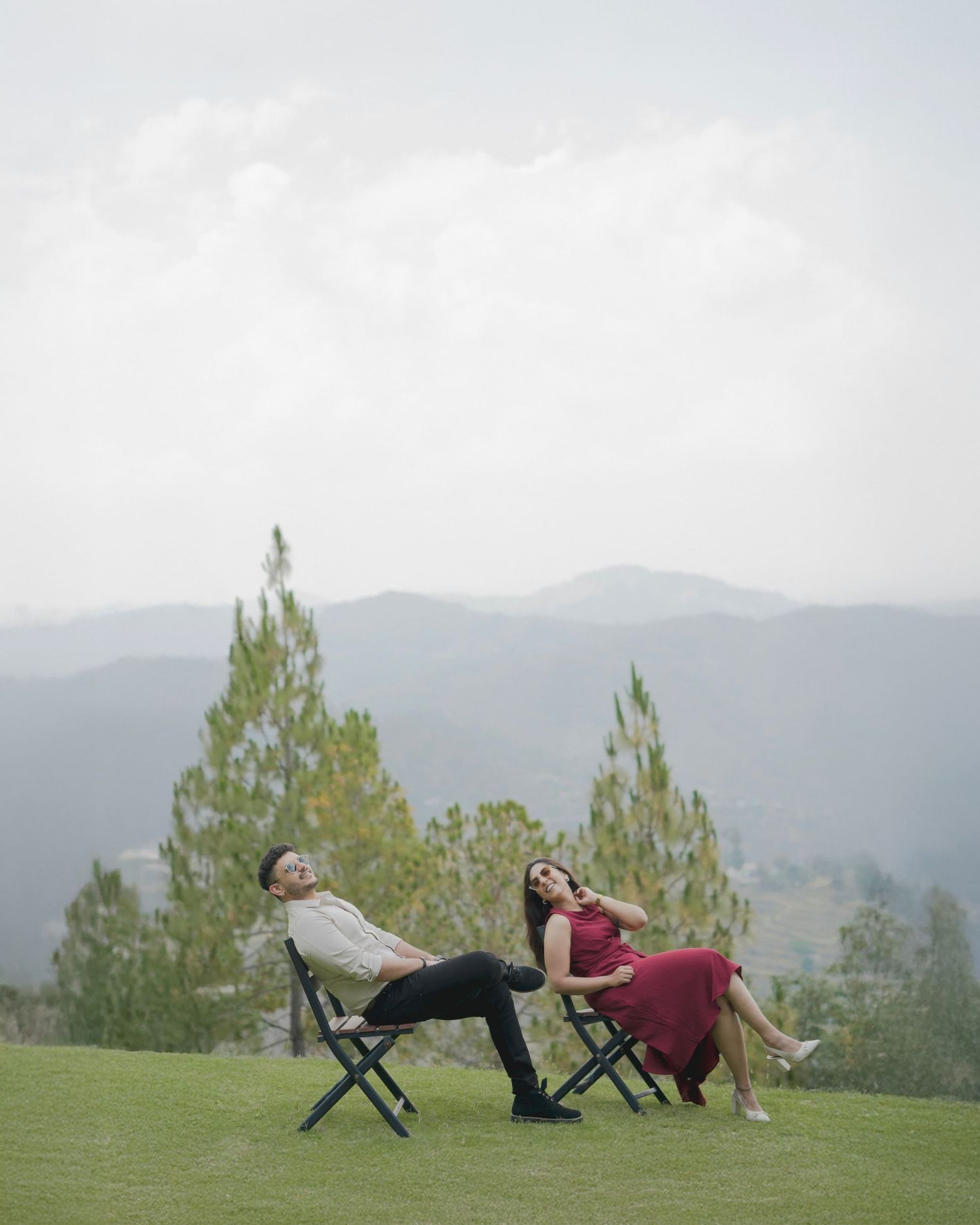 A couple relaxes in folding chairs on a green hillside with mountain and tree views under a cloudy sky.