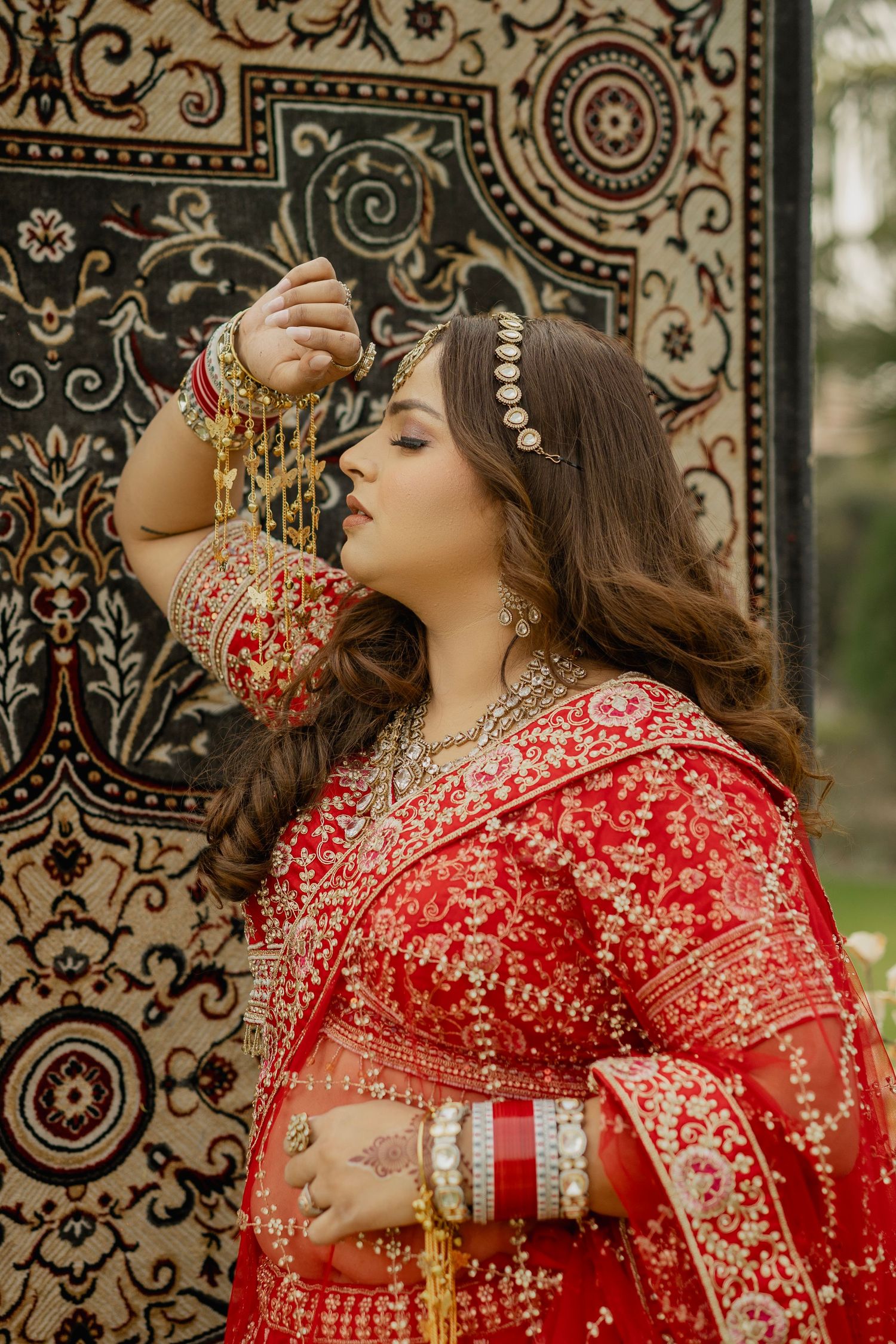 A bride in red embroidered lehenga with gold jewelry poses gracefully against an ornate dark tapestry backdrop.