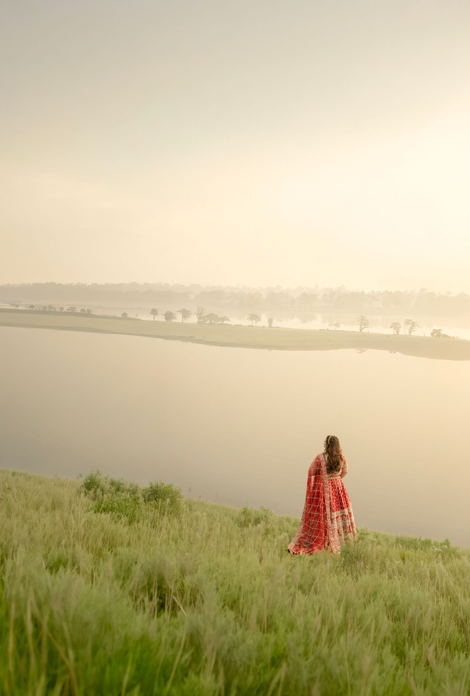 Woman in red saree standing on green hillside overlooking a misty river at golden hour.