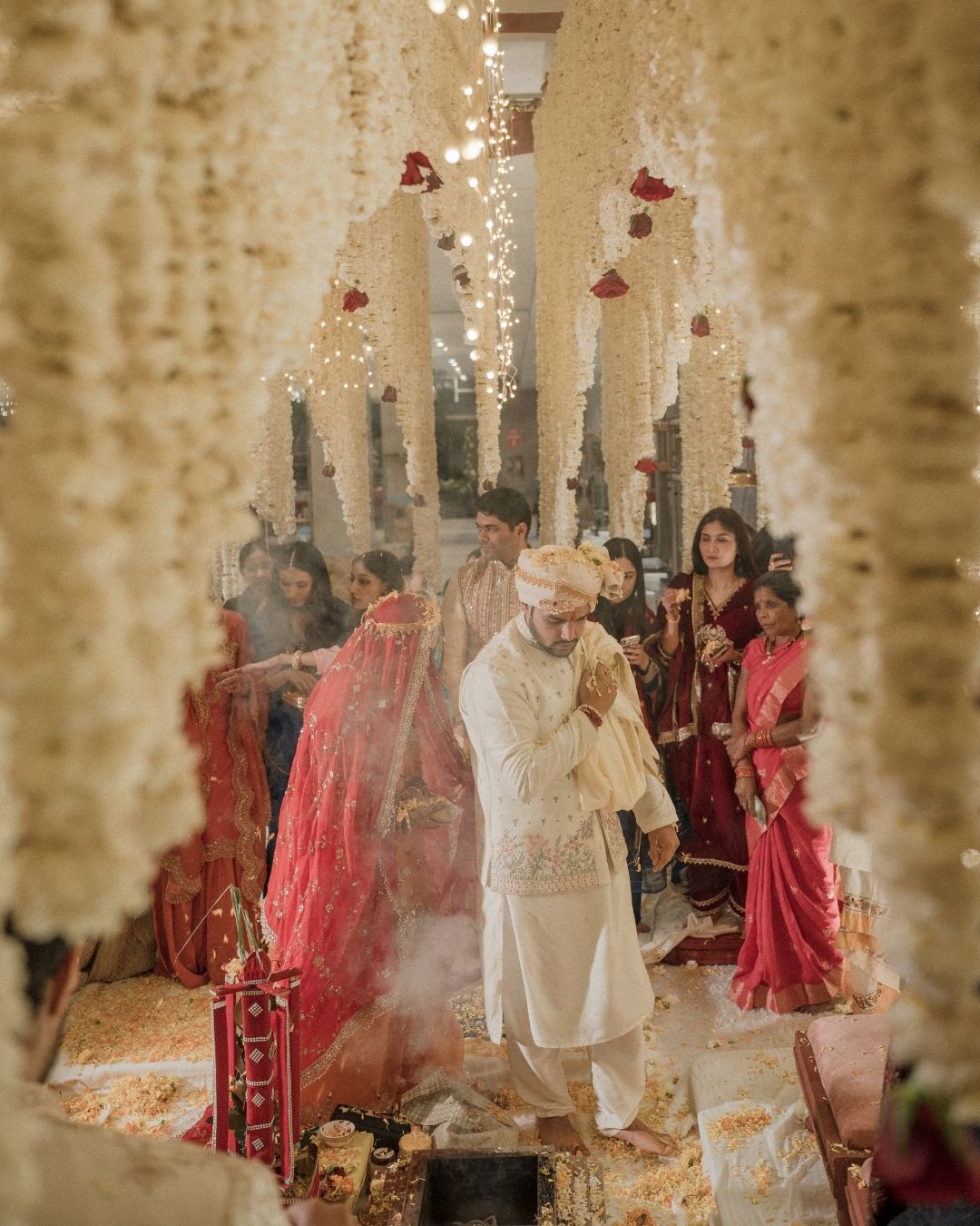 Indian bride and groom performing traditional Hindu wedding ceremony under floral decorations with guests in colorful attire.