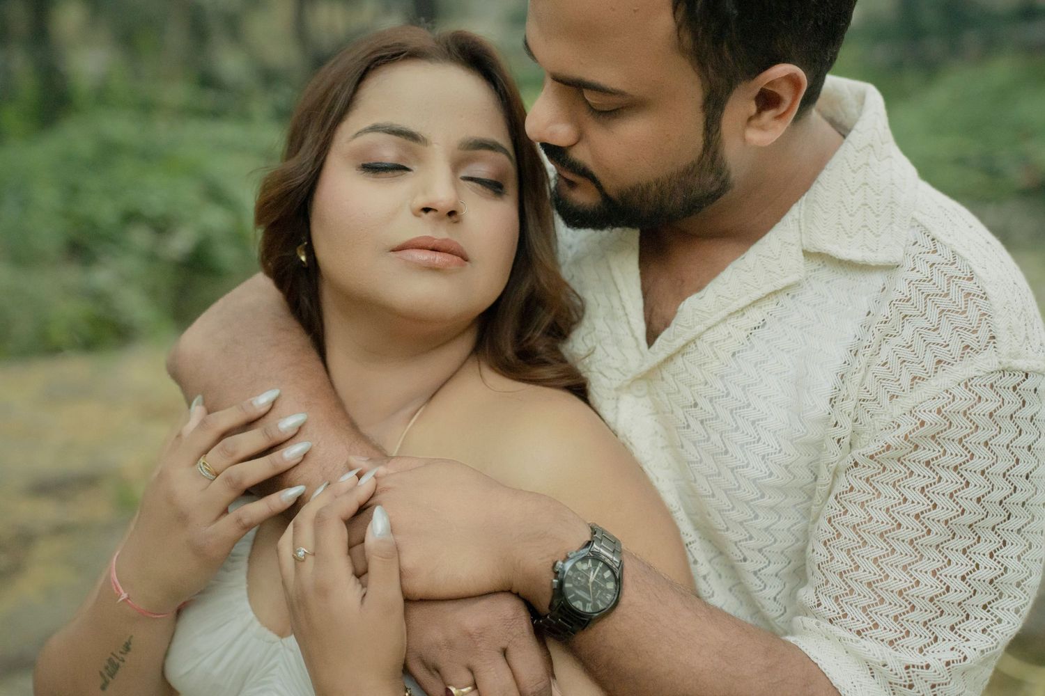 A couple in white outfits share a tender embrace outdoors, the man holding the woman from behind in a romantic pose.