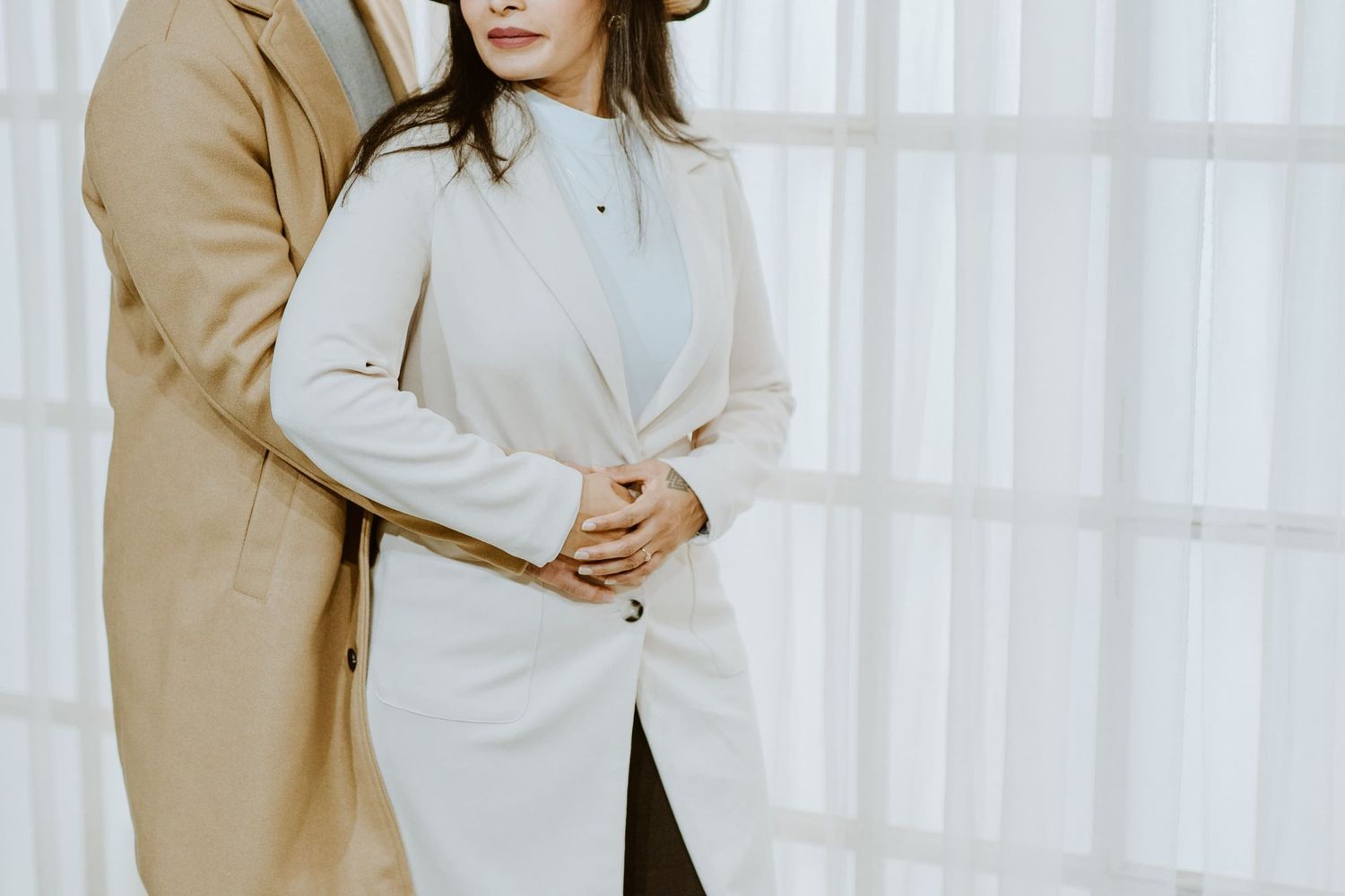 Couple in elegant beige coats standing close together in a bright, minimalist setting.
